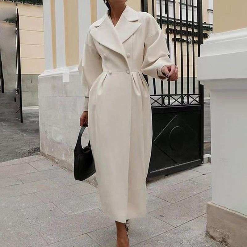 Person wearing a long beige coat holding a black handbag on a city street.