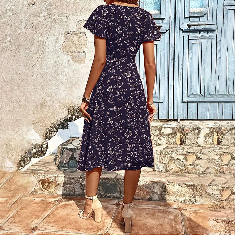 Woman wearing a dark floral dress standing on stone steps with a stone wall and wooden door in the background.