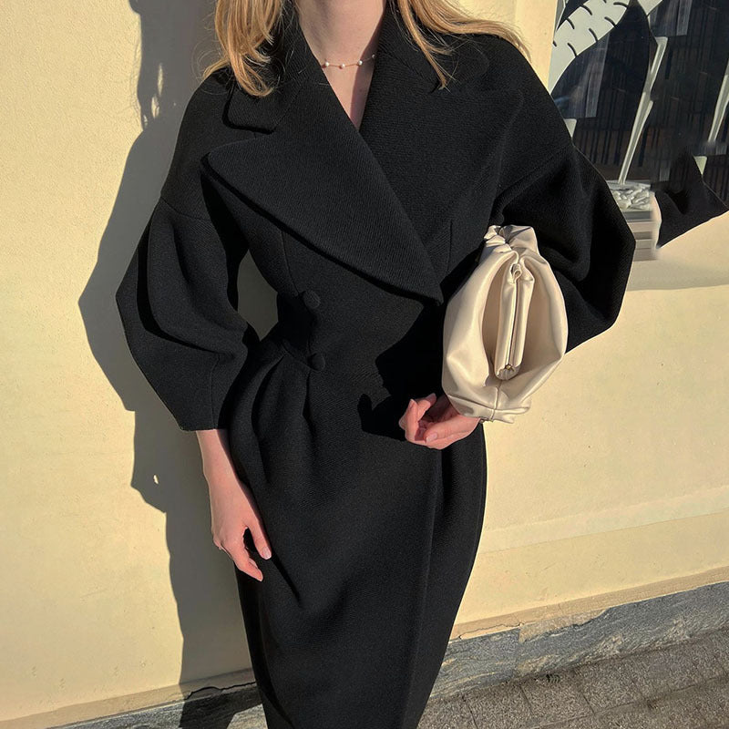 Person wearing a black coat holding a beige handbag against a light-colored wall.