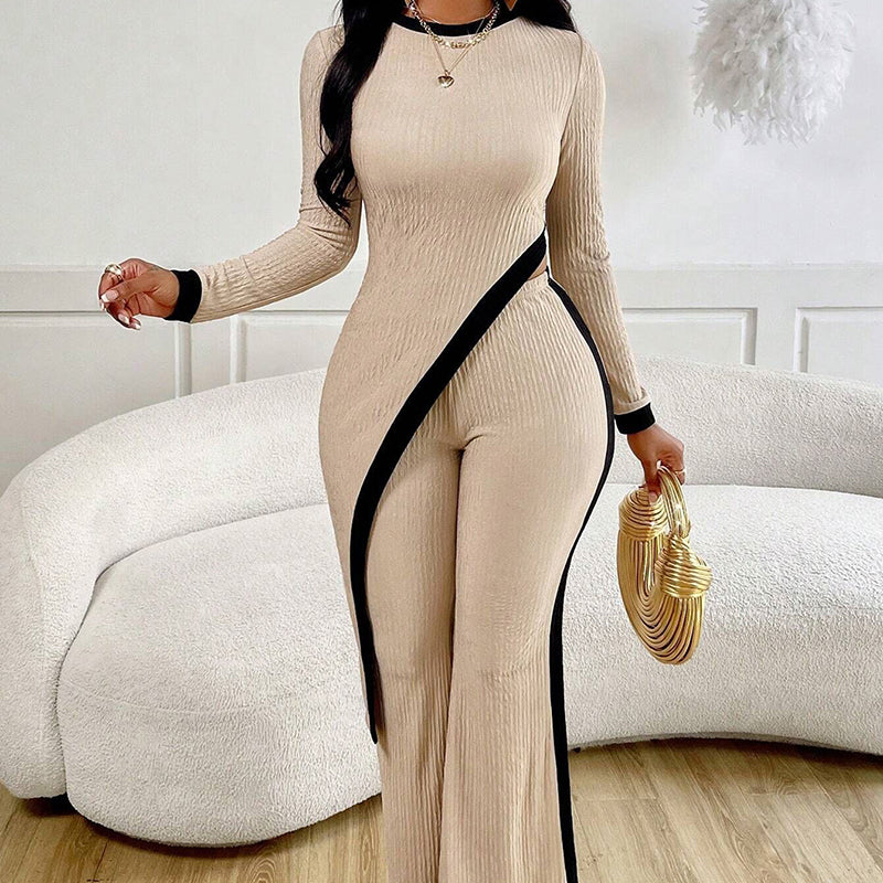 Woman wearing a beige ribbed jumpsuit with a black side stripe, holding a gold clutch in a minimalistic room.